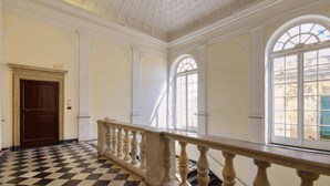 Een ruime kamer met een zwart-wit schaakbordvloer, een houten balustrade en grote ramen met gebogen bovenkanten.