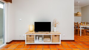 Ein Wohnzimmer mit Fernseher auf Holzständer, Korbwannen und einer Vase mit getrockneten Ästen.