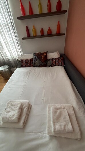 A neatly made bed with white linens and two towels, a headboard, and a shelf with decorative vases.