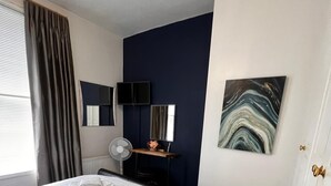 Ein Schlafzimmer mit einem Bett, einem Standventilator, einem Schreibtisch mit Spiegel und einem Bild an der Wand.