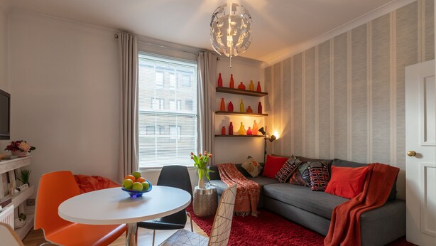 Ein modernes Wohnzimmer mit einem grauen Sofa, einer roten Decke und orangen Sesseln. Es gibt einen Esstisch mit einer Fruchtschale und einer Vase mit Blumen. Ein großes Fenster lässt viel natürliches Licht herein, und ein dekorativer Globus hängt von der Decke.