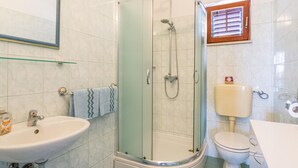 Et badeværelse med en glasvæg omkring bruseniche, en vask, et toilet og et vindue med skodder.