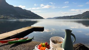 Un lac paisible, avec des montagnes en arrière-plan, un quai avec des kayaks et un coin pique-nique avec de la nourriture et une carafe.
