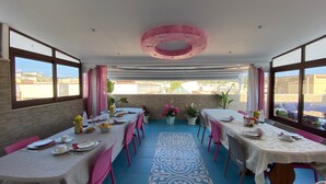 A dining area with tables set for a meal, potted plants, and a view of the cityscape through large windows. - B&B Le Onde del Mare (San Vito Lo Capo)