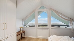 Ein Schlafzimmer mit schrägen Wänden, einem großen Fenster mit Vorhängen, einem Nachttisch mit Lampe und einem Bett mit weißer Bettwäsche.
