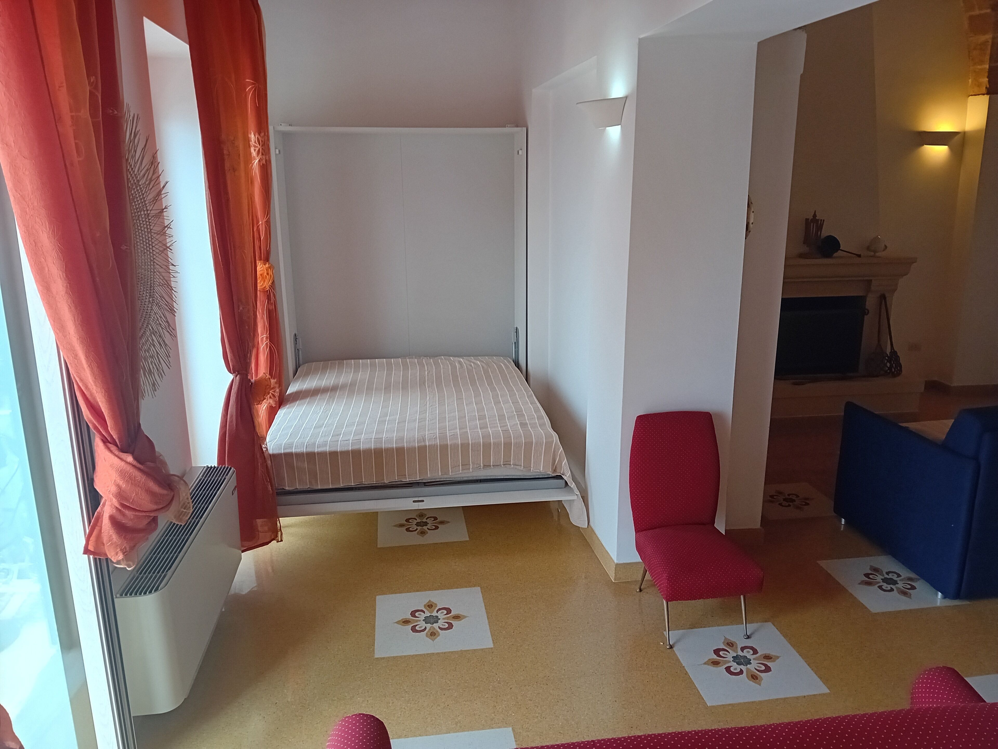 Una habitación pequeña y acogedora con una cama individual, una silla roja, una silla azul y una mesita de noche con una lámpara.