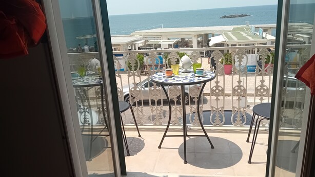 Uma varanda com vista para o mar, mesas com cadeiras e plantas em vasos.