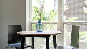 Een kamer met een ronde tafel, twee stoelen, een vaas met bloemen en een raam met uitzicht op groen.