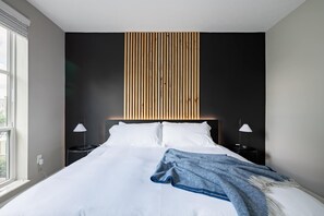 A modern bedroom with a large bed, two bedside tables, and a vertical wooden panel above the bed. - Amazing Apartment Along the River (Columbus)
