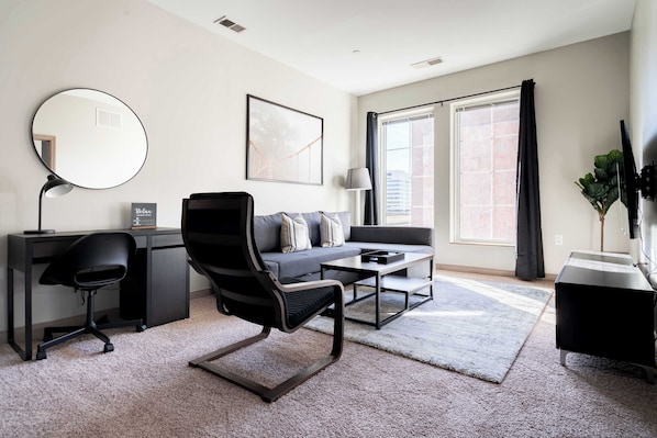 A modern living room with a sofa, a round mirror, a desk, and a large window with curtains.