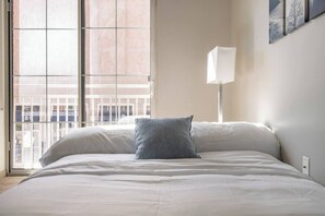 A hotel room with a large window, a bed with white bedding, a grey pillow, and a white bedside lamp. - Downtown Apt in the Heart of Columbus (Columbus)
