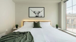 Una habitación de hotel moderna con una cama grande, cabecera, una obra de arte enmarcada de un toro, dos mesitas de noche con plantas y una ventana con vistas a la ciudad.