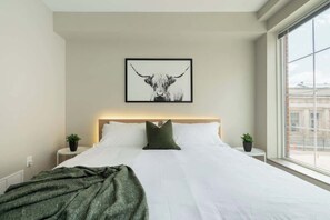 A modern hotel room with a large bed, a headboard, a framed artwork of a bull, two bedside tables with plants, and a window with a city view. - Downtown Columbus Apartment (Columbus)