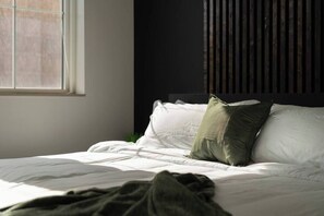 A bed with white bedding and a green pillow, a window with blinds, and a dark wooden headboard.