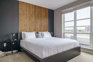 A modern bedroom with a large bed, a wooden slat headboard, bedside tables with lamps, and a large window offering a view of the outside.