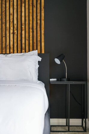 Cama bem arrumada com roupa de cama branca, mesa de cabeceira preta com abajur e painel de parede em ripas de madeira.