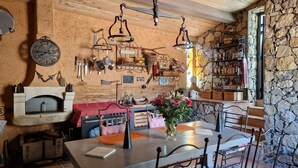 A rustic kitchen with a stone wall, wooden beams, and a variety of kitchen utensils hanging on the wall. There is a table with chairs, a vase of flowers, and a coffee machine.