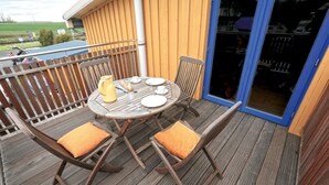 Una terraza de madera con una mesa redonda para dos personas, lista para comer, con platos, vasos y una jarra amarilla.
