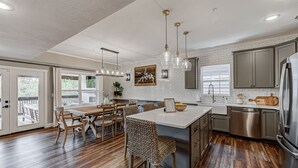 A modern kitchen with a large island, stainless steel appliances, and a dining area with wooden chairs and a white table. - Lake Access, Hot Tub, Pools, Paddleboard, Sleep 20 (Hollister)