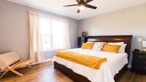 Une chambre avec un grand lit, un plancher en bois, un ventilateur de plafond, une table de chevet et une fenêtre avec des rideaux.