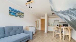 A modern living room with a blue sofa, wooden dining table with chairs, and a kitchen area with white cabinets.