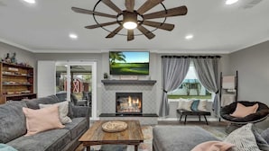 A living room with a fireplace, a ceiling fan, a television, and a view of the outdoors.