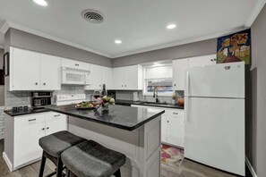 Une cuisine moderne avec des meubles blancs, un îlot central, un four à micro-ondes, un réfrigérateur et une fenêtre avec des stores.