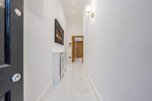 Un couloir aux murs blancs, avec un radiateur, un tableau encadré et une porte au fond.