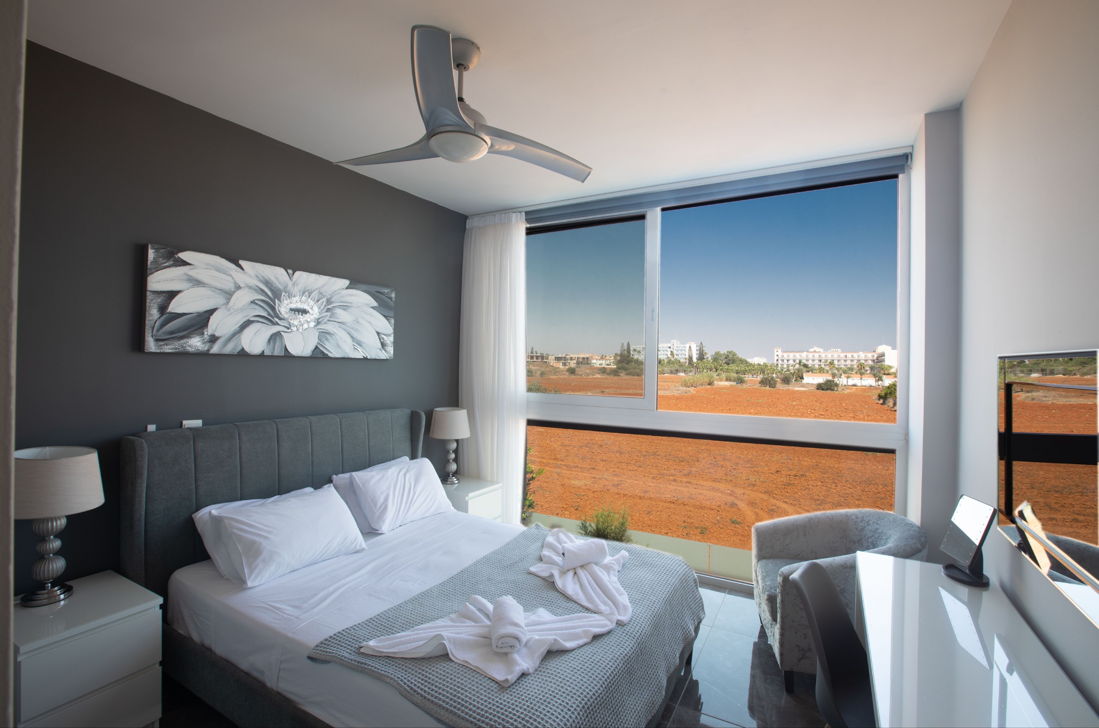 A modern hotel room with a large bed, a ceiling fan, a view of a red landscape, and a desk with a chair.