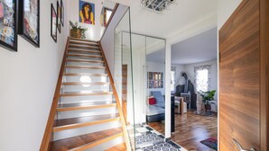 Una escalera moderna con escalones de madera y barandillas de vidrio, que conduce a una sala de estar con chimenea, estanterías y un rincón de descanso acogedor.