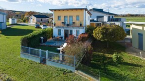 Una casa moderna de dos pisos con balcón, un patio con mobiliario exterior y un jardín cercado.