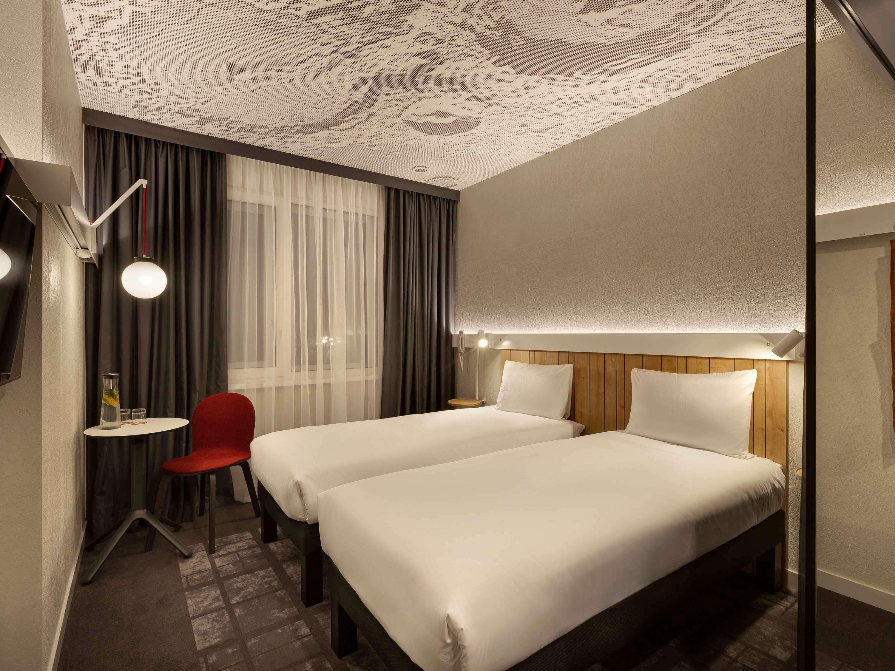 Habitación de hotel con dos camas, una mesita con una silla, una lámpara y un techo decorativo.