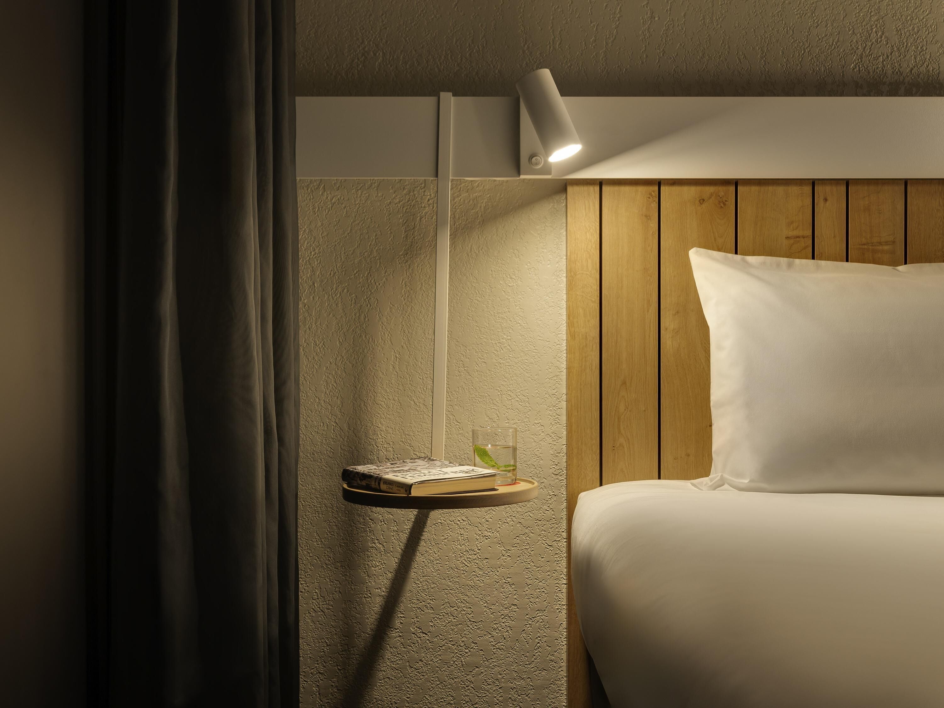 A hotel room with a bed, a bedside table with a lamp, a small shelf with a glass of water, and a curtain.
