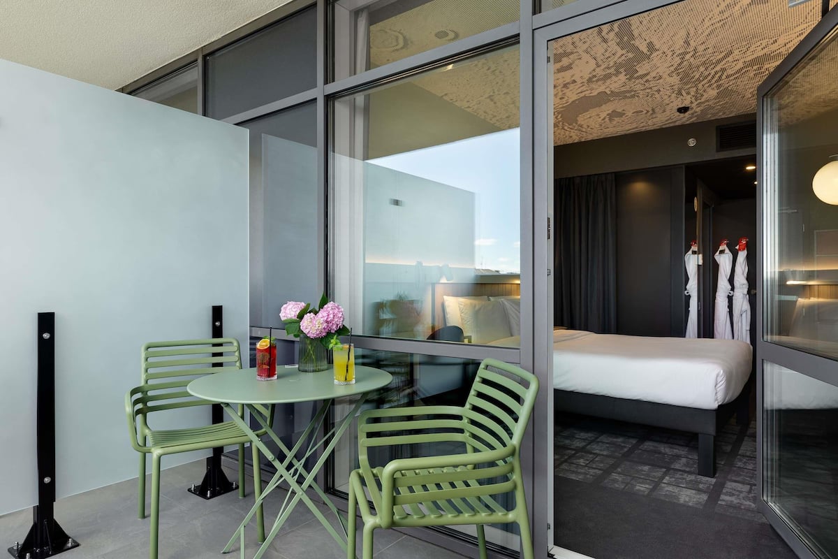 Ein modernes Hotelzimmer mit einer Glaswand, einem Bett, einem kleinen Tisch mit zwei Stühlen und einer Vase mit Blumen.