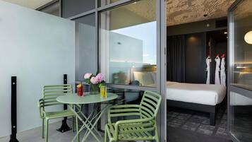 A modern hotel room with a glass wall, a bed, a small table with two chairs, and a vase with flowers.
