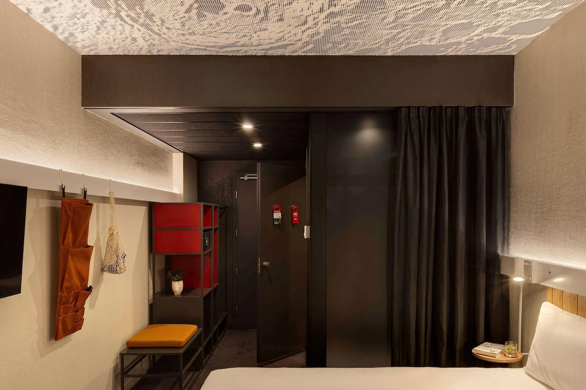 Ein modernes Hotelzimmer mit einem großen Bett, einer kleinen Sitzecke, einem roten Regalelement und einer dunklen Tür.