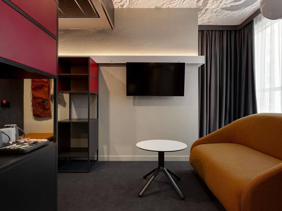 Una habitación de hotel moderna con una televisión de pantalla plana montada en la pared, una pequeña mesa redonda, un sofá y un armario empotrado con secciones rojas y negras.