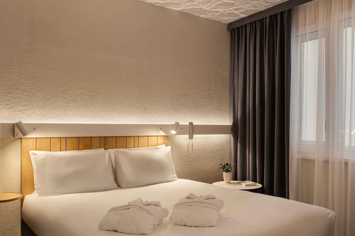 Ein Hotelzimmer mit einem ordentlich bezogenen Bett, einem Nachttisch mit Lampe und einem Fenster mit Vorhängen.