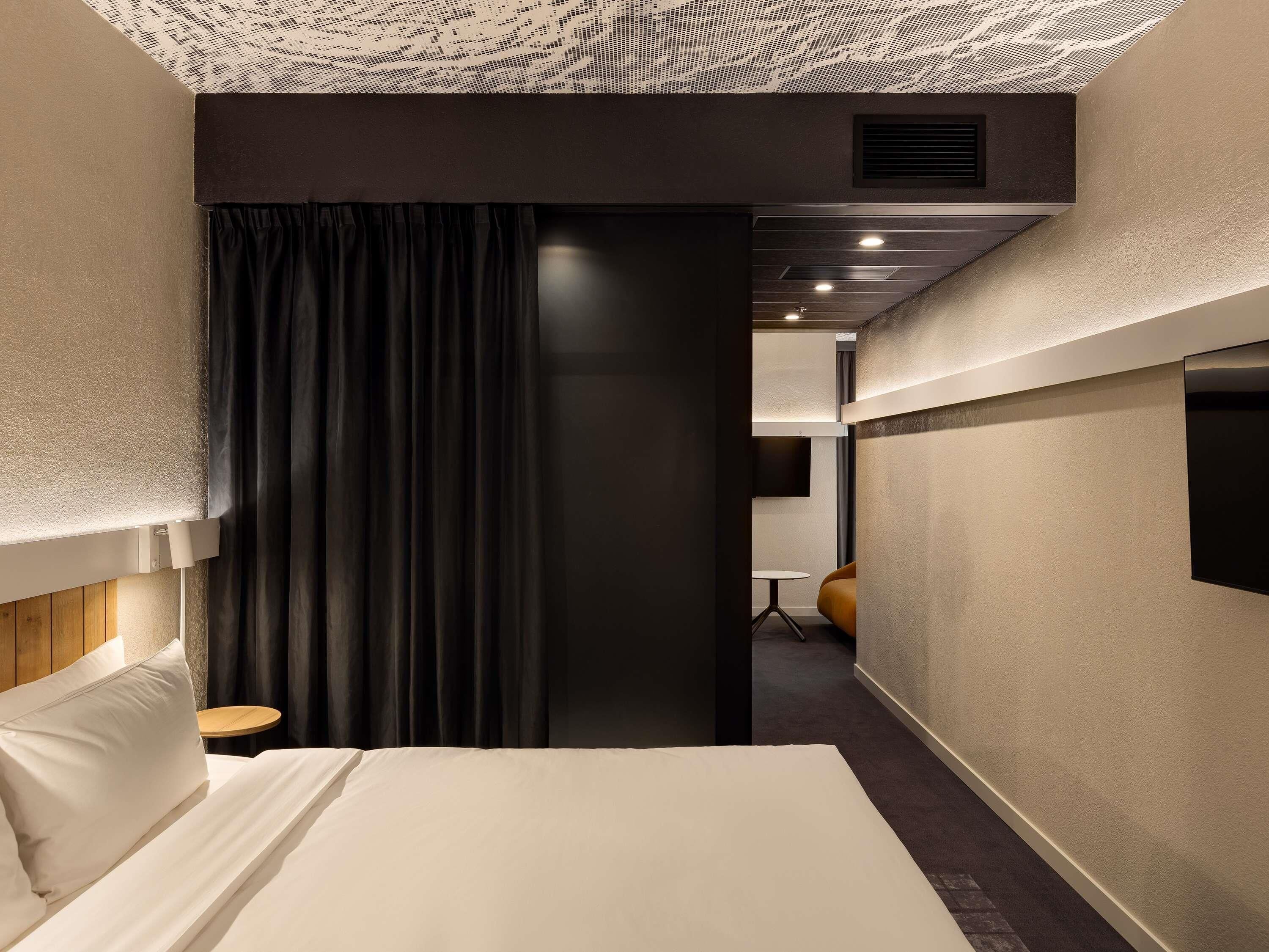 A hotel room with a bed, a small table, a chair, a TV, and a dark curtain.