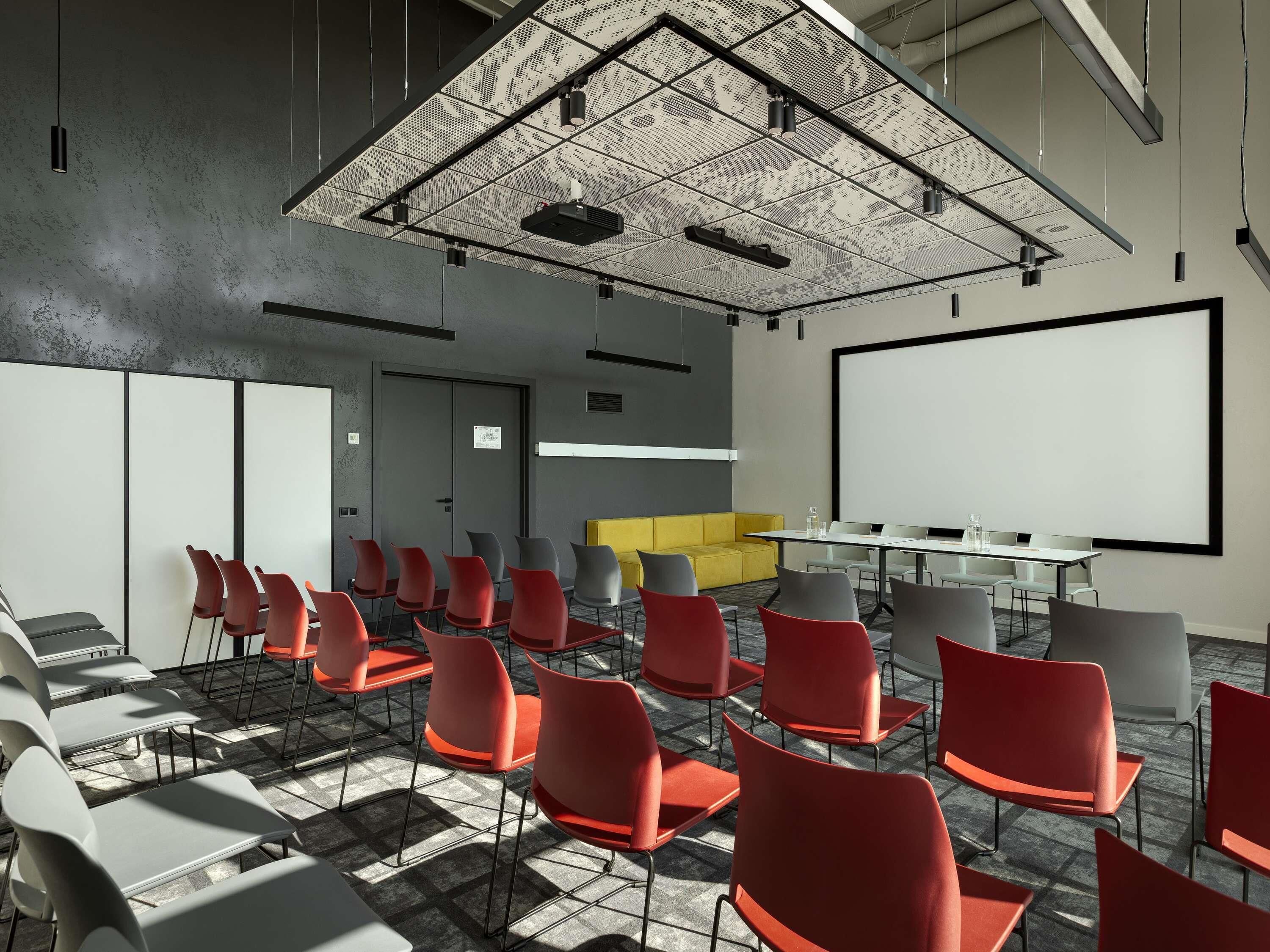 A conference room with a large white screen, red and grey chairs, a yellow sofa, and a ceiling with a grid pattern.