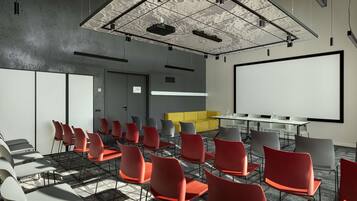 A conference room with a large white screen, red and grey chairs, a yellow sofa, and a ceiling with a grid pattern.