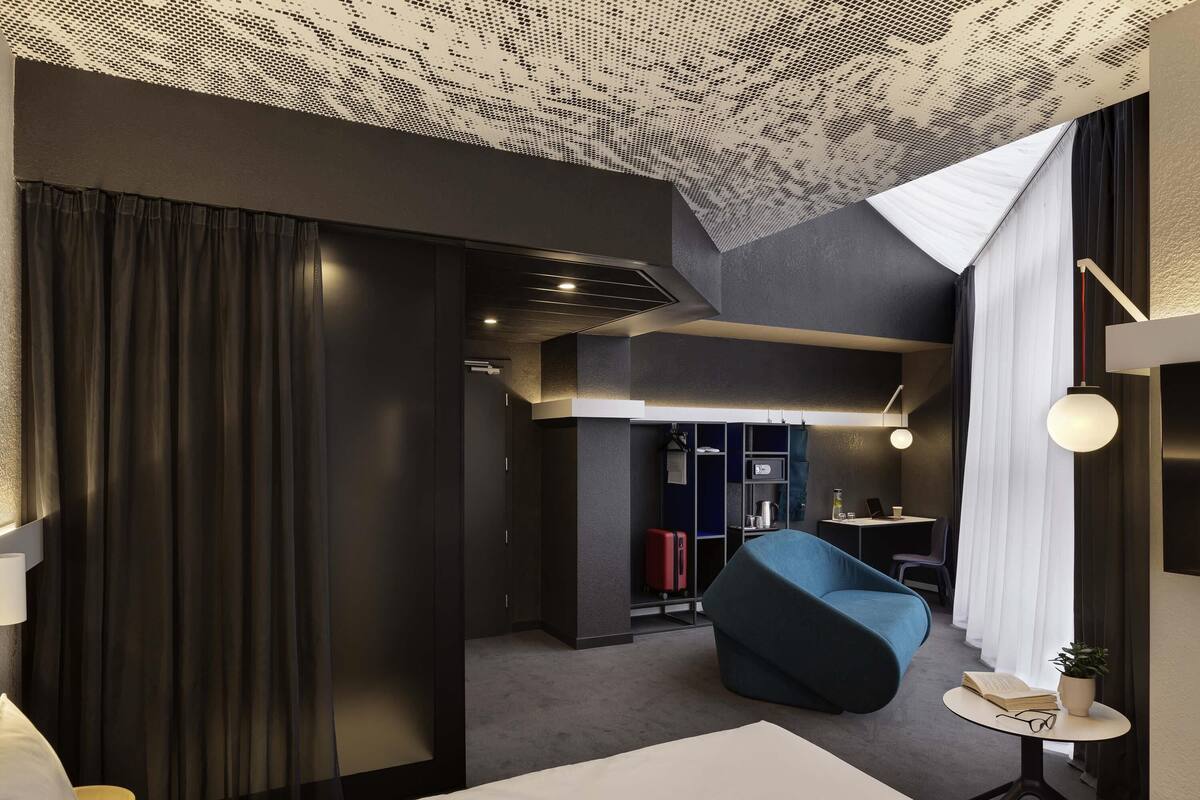 A modern hotel room with a bed, a desk, a chair, a small table with a plant, and a kitchenette area.