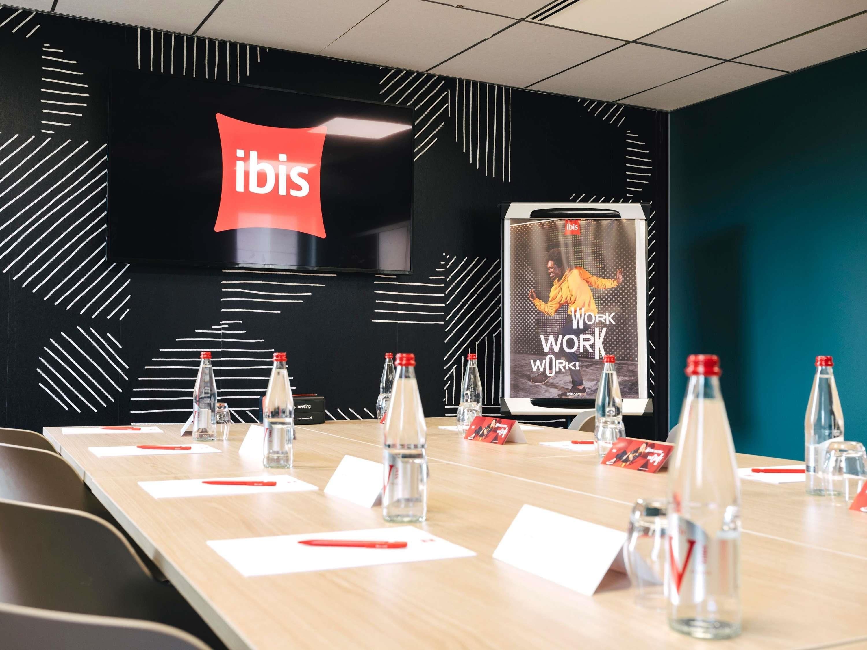 A conference room with a long table, chairs, water bottles, name cards, and a screen displaying 'ibis WORK WORK WORK!'