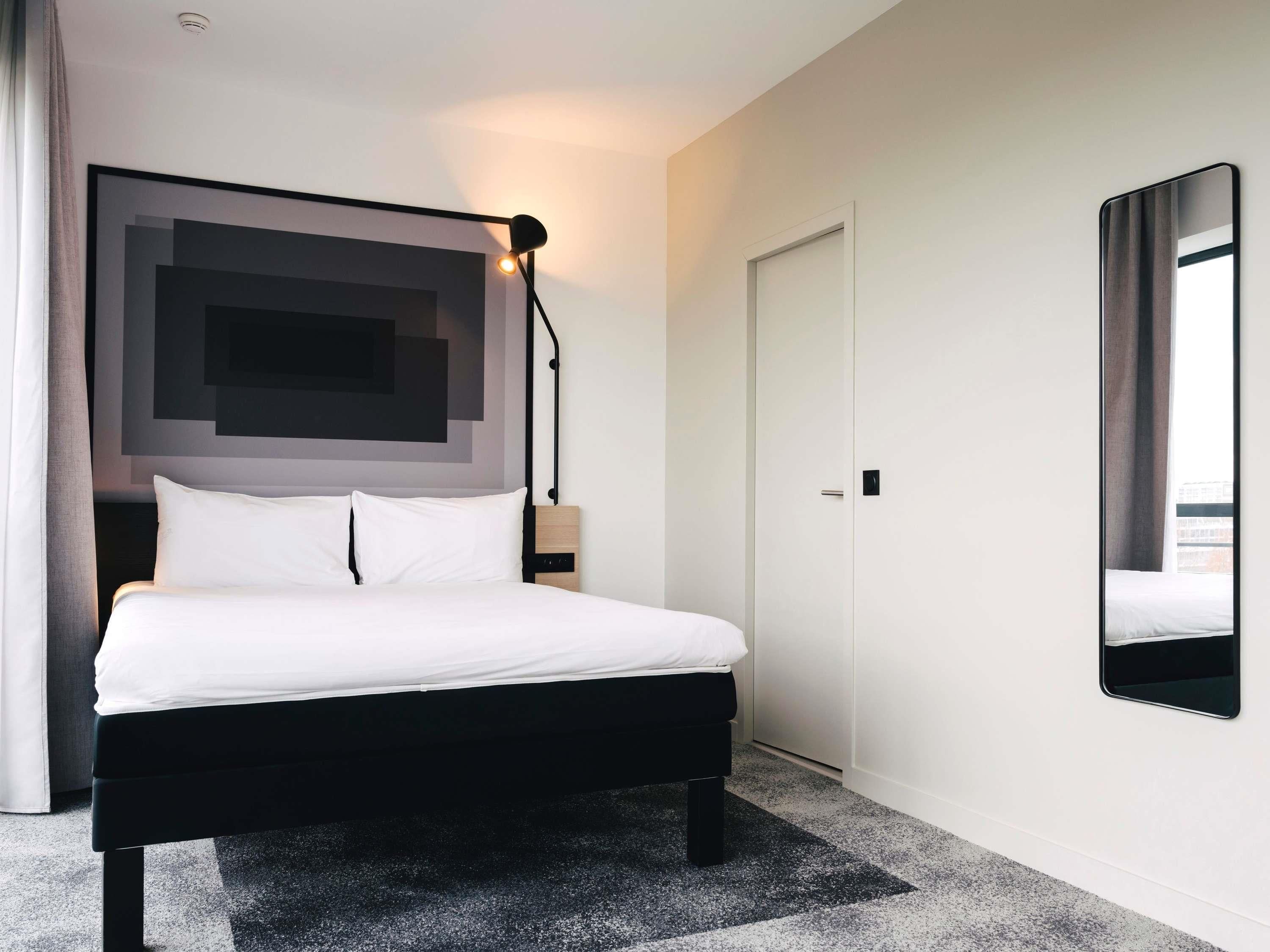 A modern hotel room with a large bed, a bedside table, a floor lamp, and a large mirror.