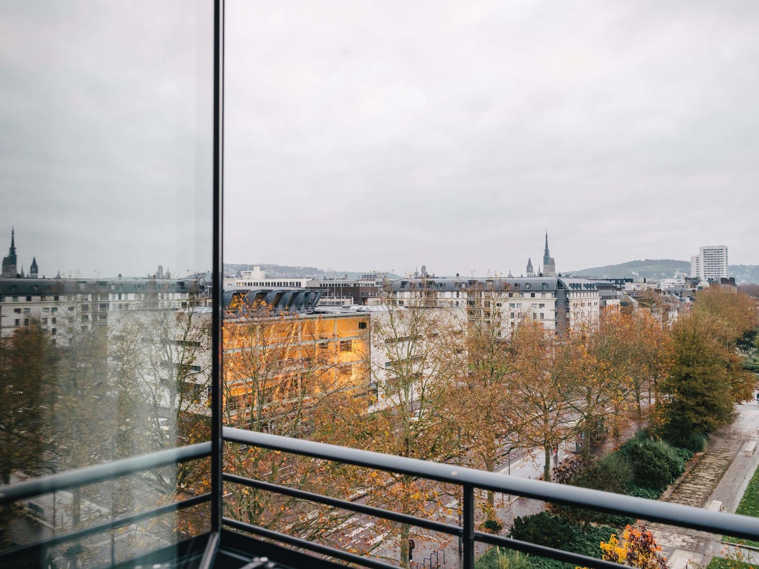 Stadsgezicht vanuit een hoog gebouw met een glazen raam, met herfstbomen en verschillende gebouwen.