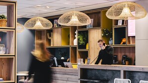 Zona de recepción con estanterías de madera, lámparas colgantes de mimbre y un mostrador con teléfono y ordenador portátil.