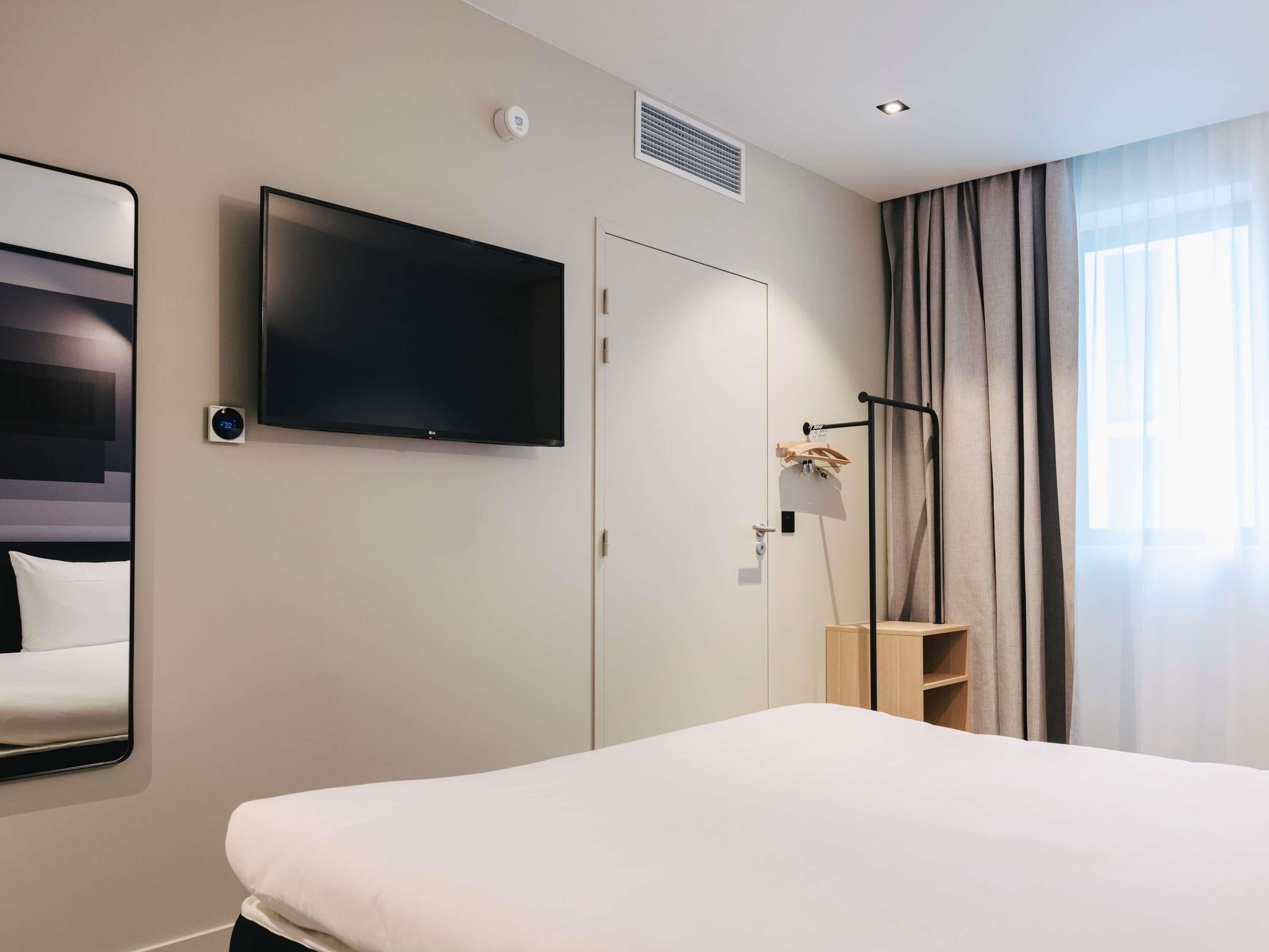 A hotel room with a bed, a television mounted on the wall, a door, a window with curtains, and a small shelf.