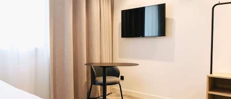 A hotel room with a bed, a round table with a chair, a TV mounted on the wall, and a bookshelf.