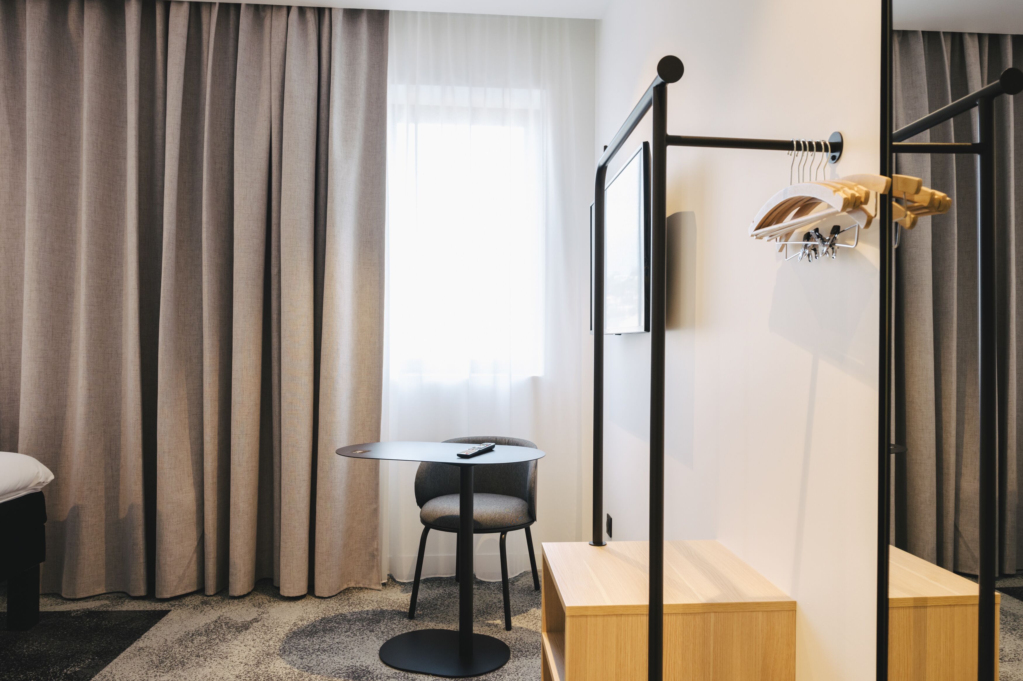 Ein modernes Hotelzimmer mit einem Bett, einem kleinen runden Tisch mit Stuhl, einer hölzernen Bank, einem Kleiderständer und einem großen Fenster mit Vorhängen.