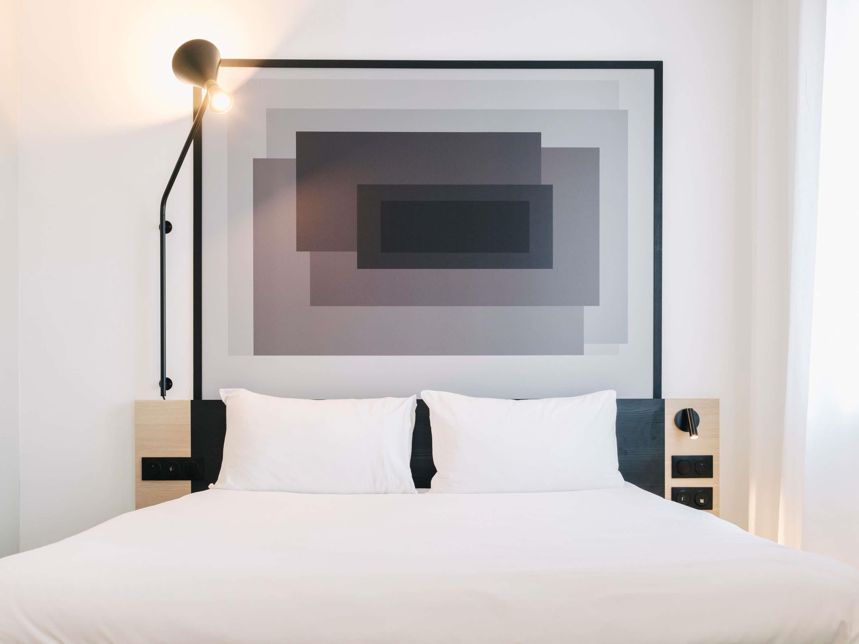Una habitación de hotel moderna con un diseño minimalista, que incluye una cama grande con ropa de cama blanca, un cabecero negro y una obra de arte mural geométrica.